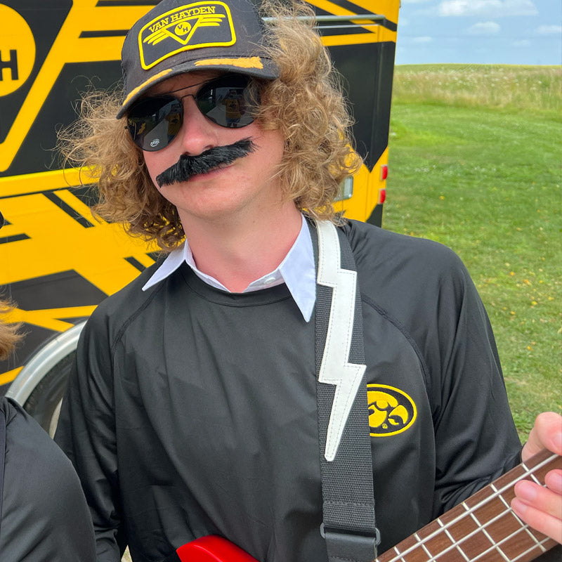 Mike Van Hayden wearing a trucker hat and sunglasses, holding a bass guitar, with a Van Halen branded vehicle in the background.