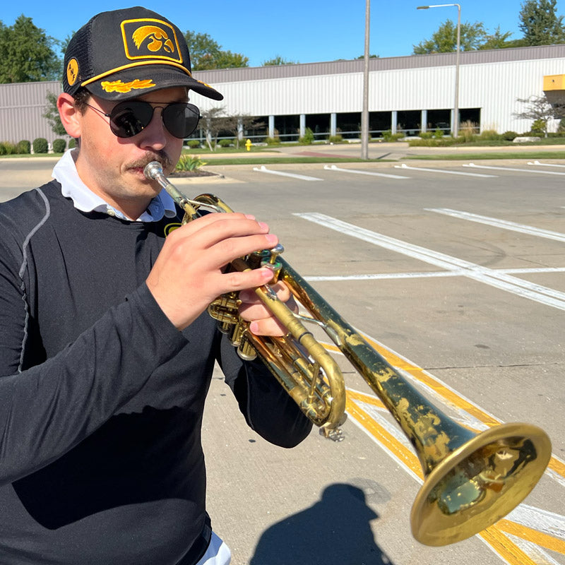 Montrose Van Hayden playing a trumpet in a parking lot with a building and trees in the background